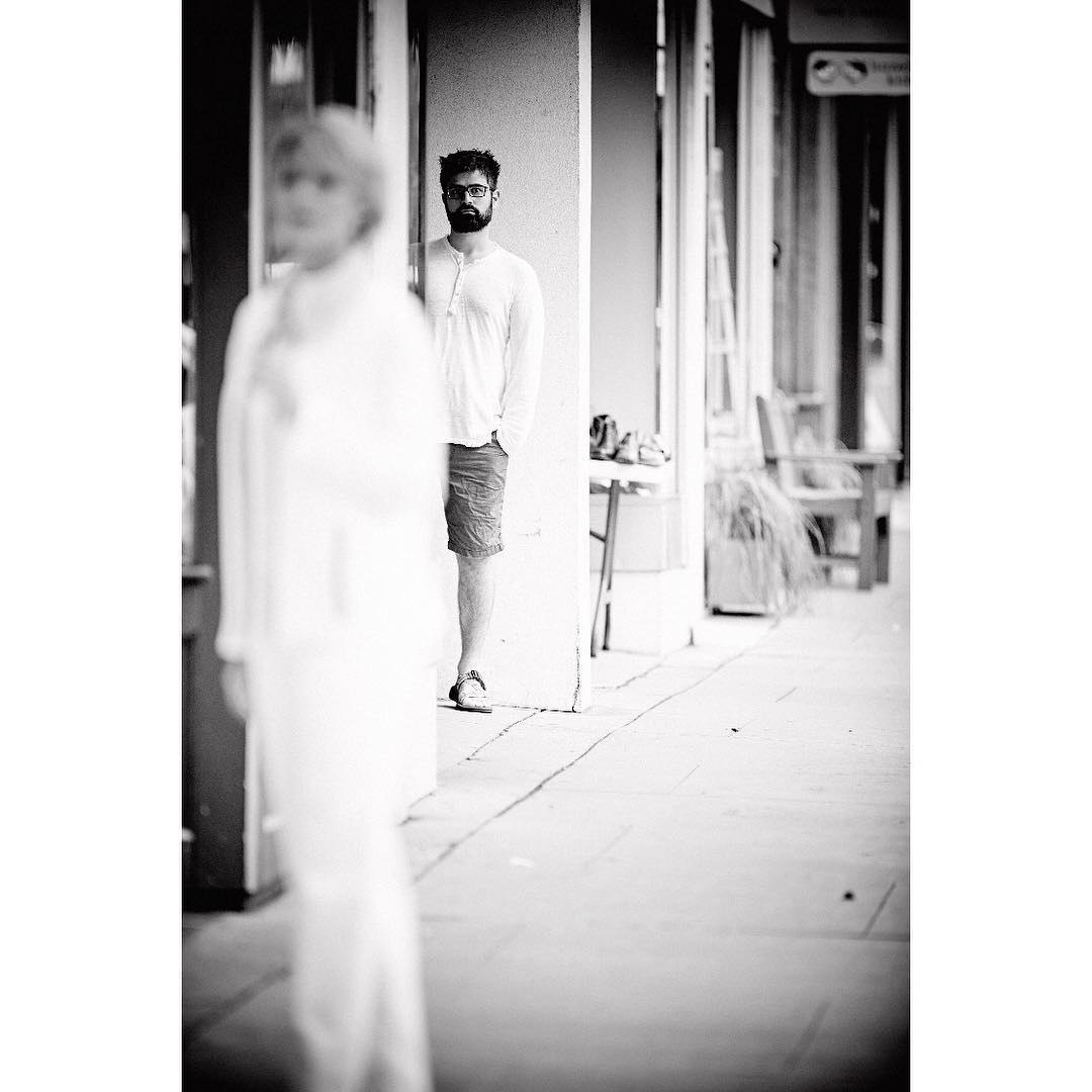 Black and white shot of a man with a beard standing outside, partially hidden behind a white column, with a blurred person in foreground.