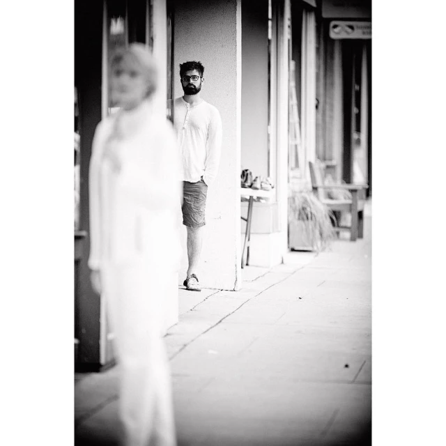 Black and white shot of a man with a beard standing outside, partially hidden behind a white column, with a blurred person in foreground.