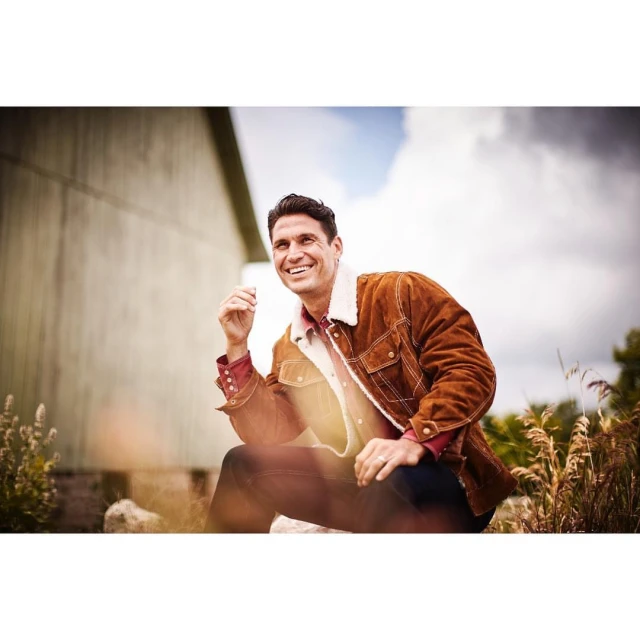 Smiling man sits outdoors wearing a tan shearling jacket and red shirt. He is surrounded by weeds and a barn is in the background.
