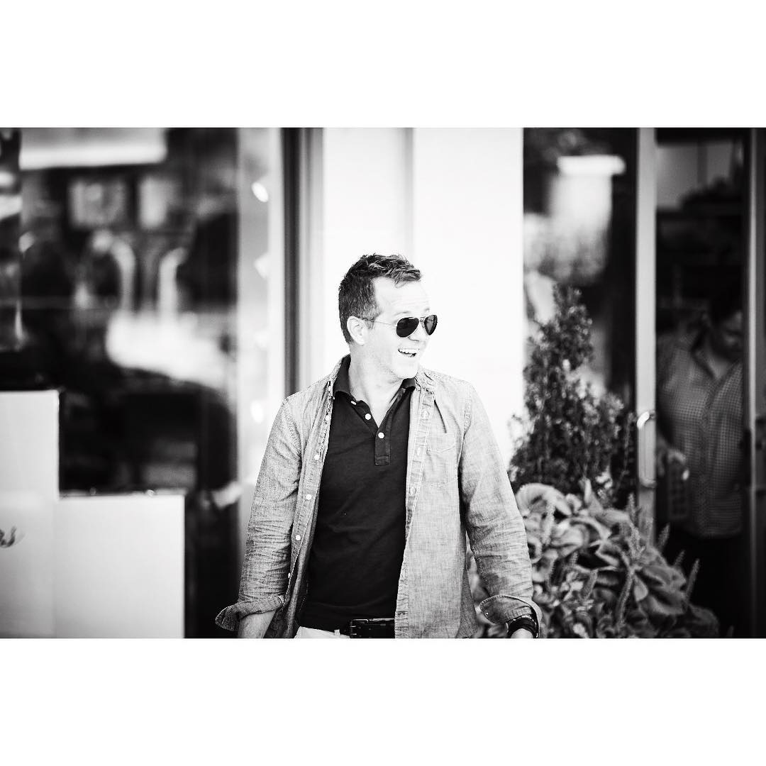 Monochrome portrait of a smiling man in sunglasses and casual shirt, standing outdoors near a window with reflections.