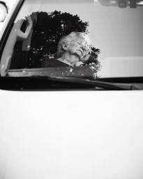 Monochrome portrait of an elderly woman sleeping inside a car, viewed through the windshield with tree reflections.