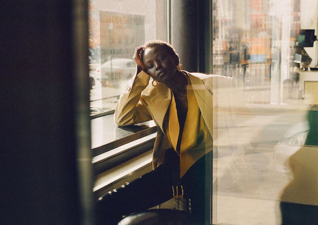 Woman in yellow blazer and black pants poses indoors, resting her head on her hand, with a city street scene visible through a window.