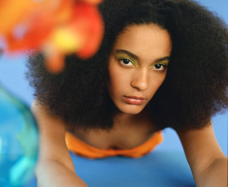Close-up of a young Black woman with afro hairstyle and green eyeshadow looking directly at the camera in a studio environment.