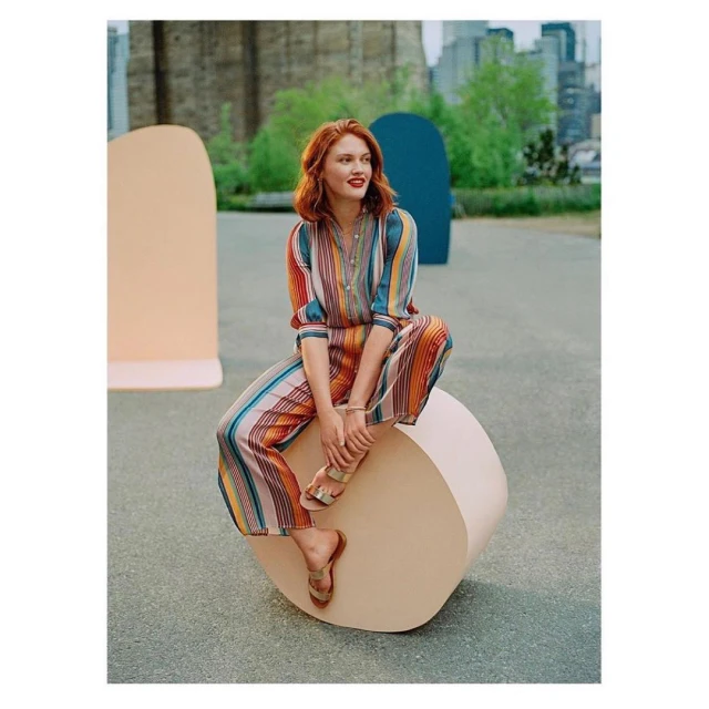 Redhead woman wearing a striped jumpsuit sitting on a rounded prop, with urban buildings visible in the background, captured in daylight.
