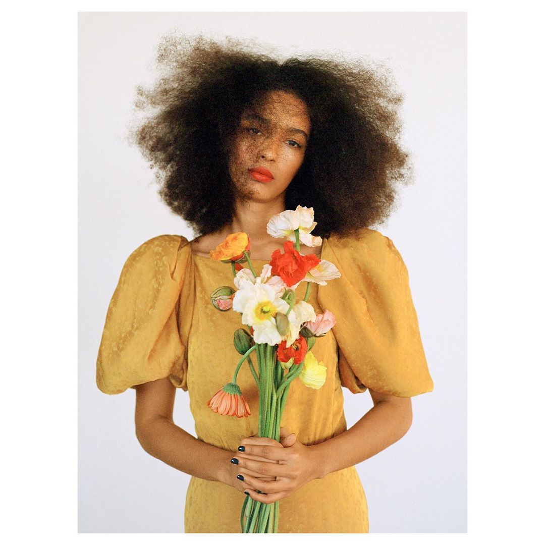 A woman with an afro hairstyle poses indoors wearing a yellow dress, holding a bouquet of flowers in a portrait-style photograph.