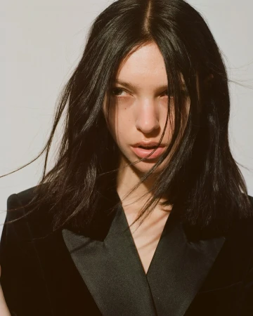 Portrait of a woman with dark hair wearing a black blazer against a light wall by Tiffany Nicholson