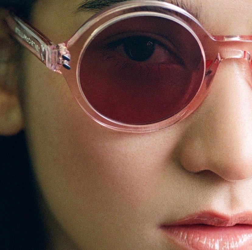 Close-up studio shot of a woman's face wearing pink-rimmed sunglasses with dark red lenses, showcasing beauty and fashion.