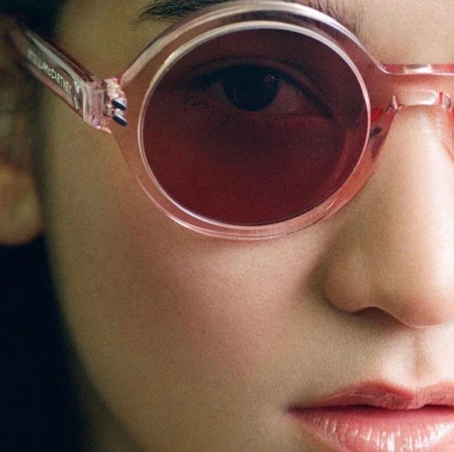 Close-up studio shot of a woman's face wearing pink-rimmed sunglasses with dark red lenses, showcasing beauty and fashion.