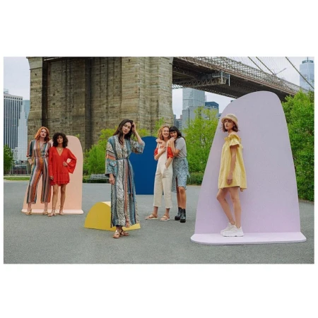 Five women pose in dresses and casual wear near colorful geometric shapes with the Brooklyn Bridge and NYC buildings in the background.