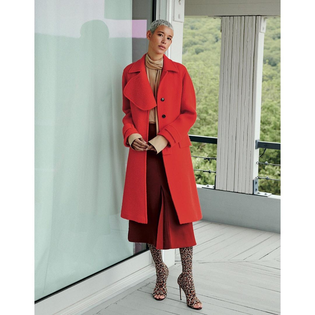 A fair-skinned adult woman with short gray hair poses indoors wearing a red wool coat, maroon skirt, patterned hosiery, and open-toe high heels.