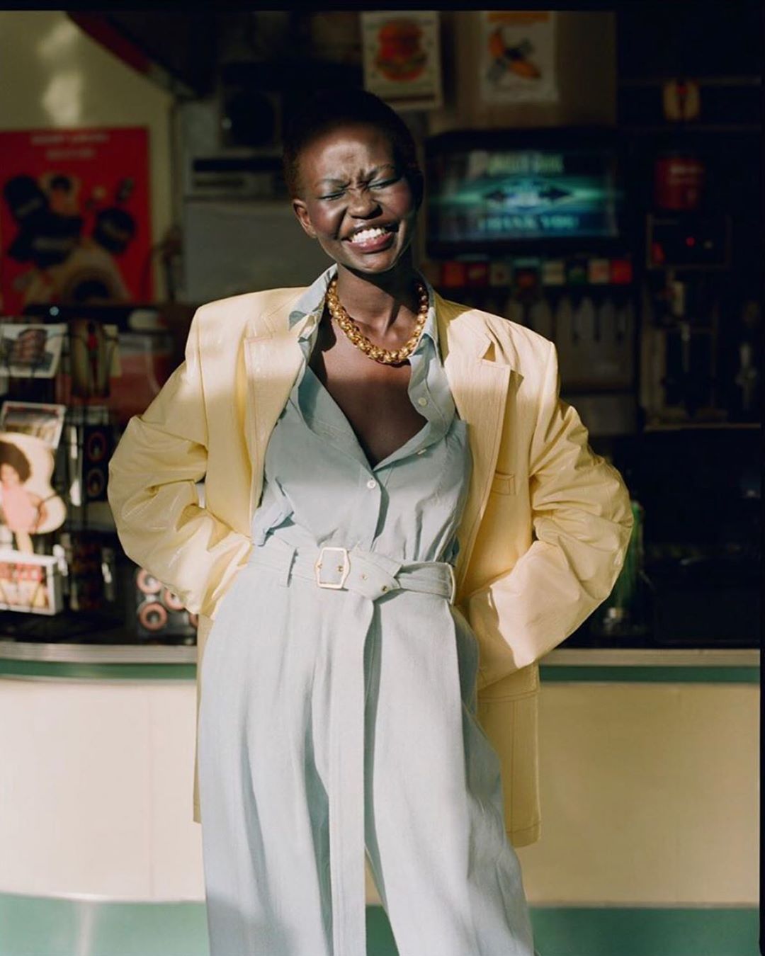 Smiling Black woman in blue jumpsuit and yellow blazer poses in front of a diner counter with vintage posters and equipment.