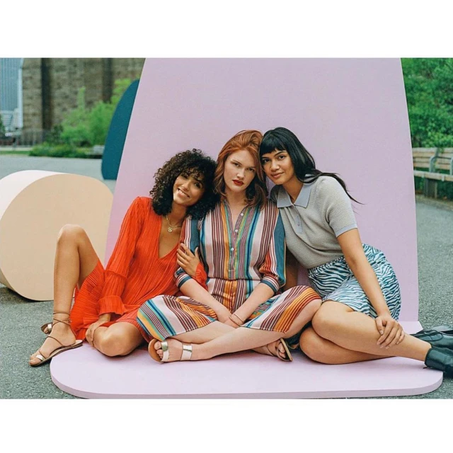 Three diverse women model colorful dresses and skirts while sitting together against a pastel backdrop in an outdoor setting.