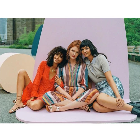 Three diverse women model colorful dresses and skirts while sitting together against a pastel backdrop in an outdoor setting.