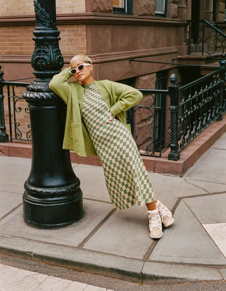 Woman in a green checkered dress and cardigan leans against a black lamppost in an urban setting, accessorized with round sunglasses and beige boots.