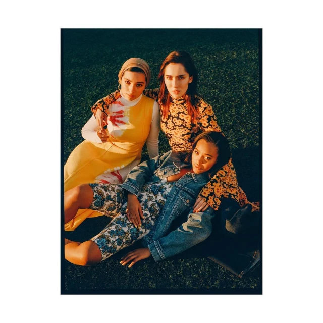Three young women are sitting close together on a lawn wearing stylish retro clothing in a medium shot with natural lighting.