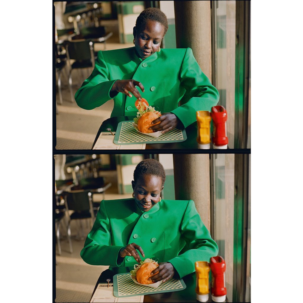 Woman in a green suit jacket sits at a diner table preparing to eat a burger with condiments, inside a retro-style restaurant.