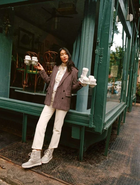 Stylish woman in a herringbone blazer stands outside a green-framed building, holding trays of coffee in a casual, outdoor scene.