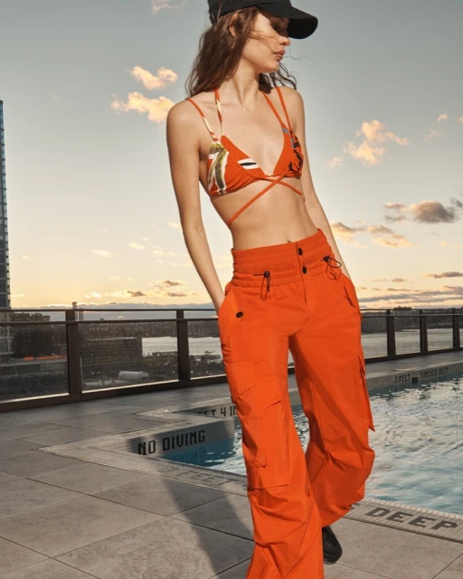 Woman wearing orange pants and bikini top poses on rooftop with city skyline and swimming pool under a partly cloudy sky at daytime.