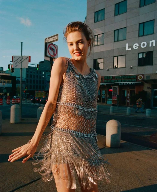 A model in a silver fringe dress poses on a city street with buildings, street signs, and traffic lights visible in the background.