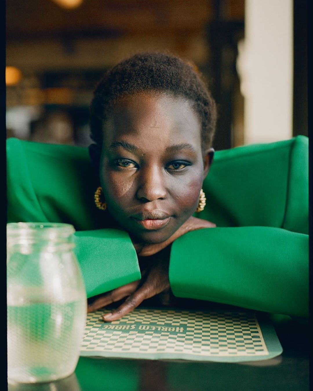 Close-up portrait of a woman in a green blazer resting her arms on a table with a drink and a placemat featuring 'Harlem Shake' text.