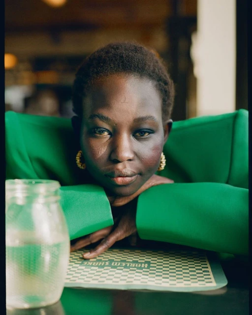 Close-up portrait of a woman in a green blazer resting her arms on a table with a drink and a placemat featuring 'Harlem Shake' text.