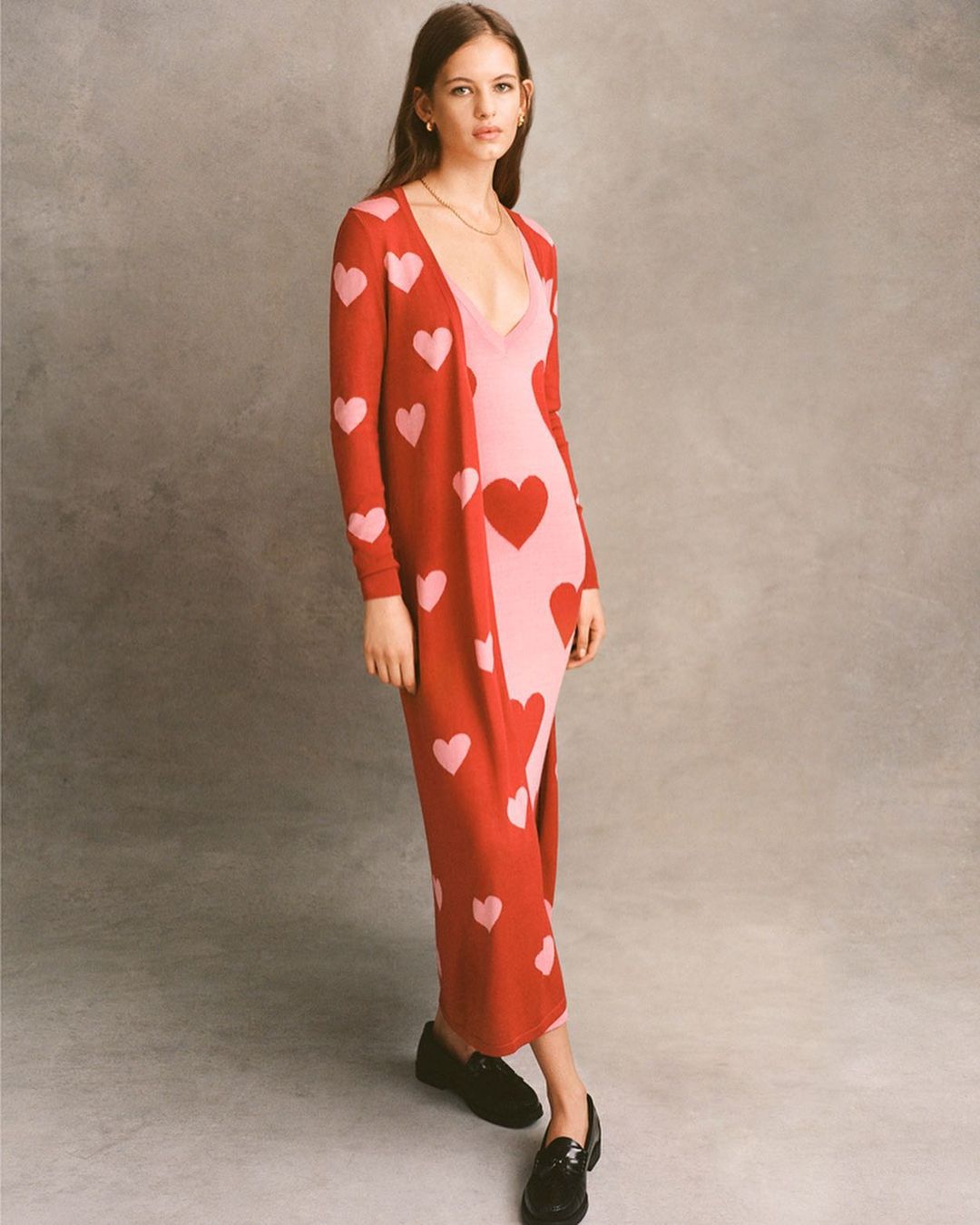 A young woman in a pink dress and a red cardigan with pink hearts stands in front of a neutral-colored background in a studio.