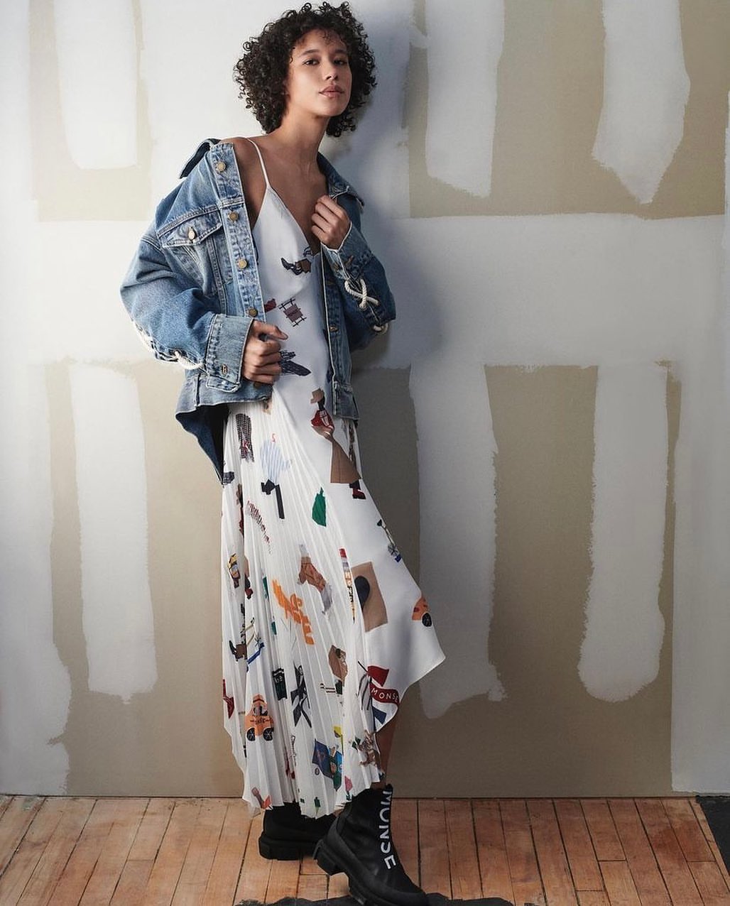 A young adult woman poses in a printed white dress with a denim jacket and black boots against a wall in a studio setting.