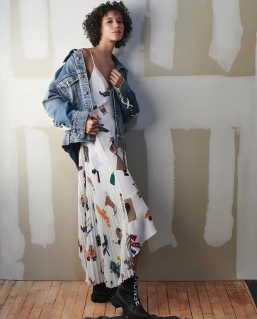 A young adult woman poses in a printed white dress with a denim jacket and black boots against a wall in a studio setting.