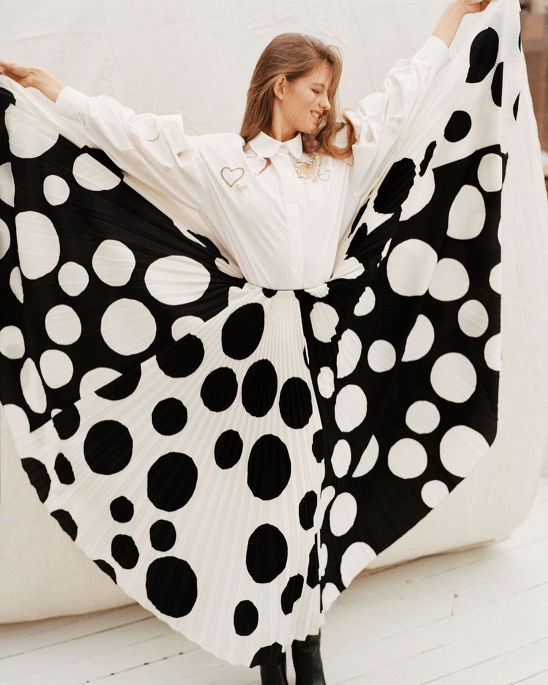 A stylish young woman wearing a white blouse and black and white polka dot skirt poses with arms raised in a light-filled setting.
