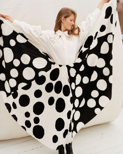 A stylish young woman wearing a white blouse and black and white polka dot skirt poses with arms raised in a light-filled setting.