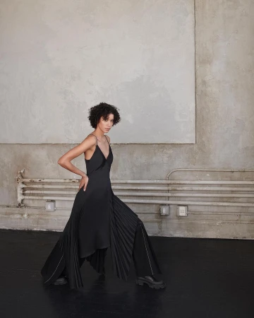 Model with curly hair wearing a black slip dress and wide trousers posing against a weathered industrial concrete wall