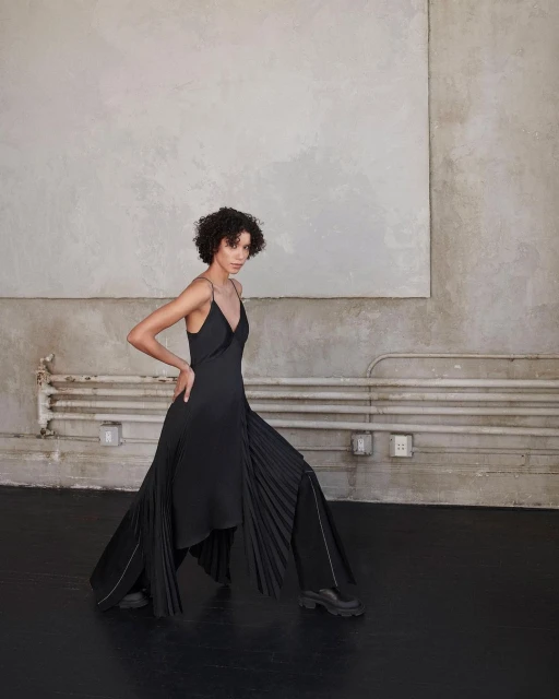 Fashion shot of an adult woman in black dress and dark boots posing in front of a concrete wall with industrial pipe feature.