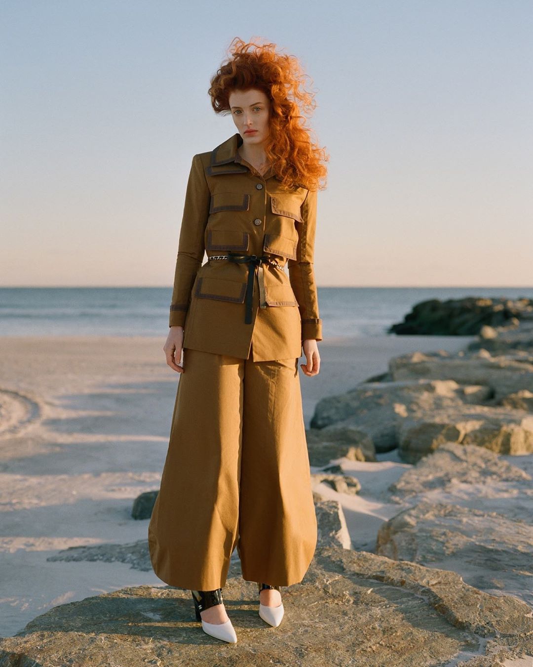 Red-haired woman in a tan outfit stands on a beach rock formation with ocean and sky in the background on a sunny day.