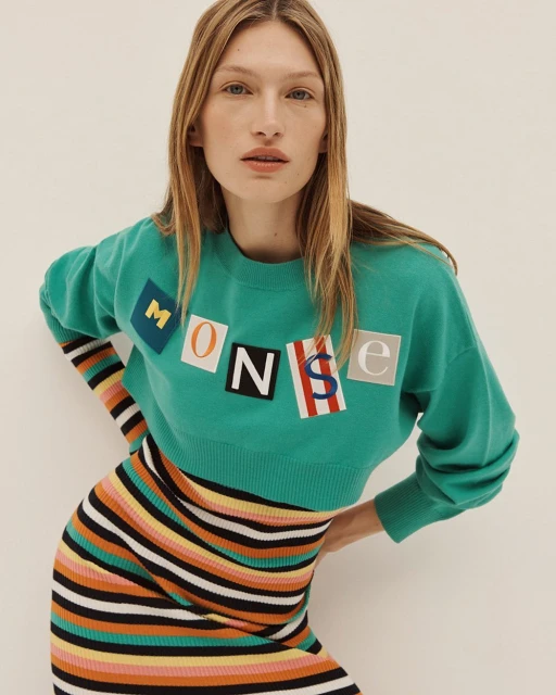 Adult female fashion model poses in a green sweater with letter graphics and a striped skirt against a neutral backdrop.