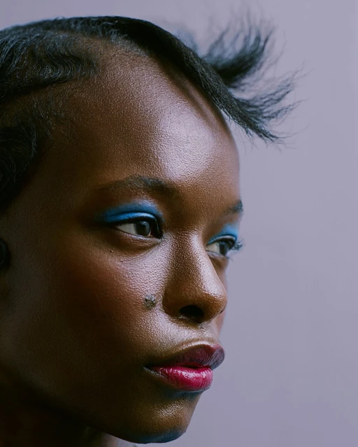 Close-up portrait of an African American woman with vibrant blue eyeshadow and red lipstick on a pale lavender background.