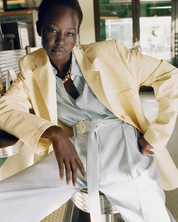 Model wearing a pale yellow blazer and matching belted trousers, seated indoors on a round stool.