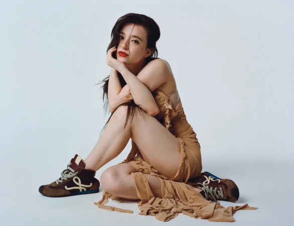 A seated woman with dark hair and red lipstick wearing a beige draped dress and brown sneakers against a white background.