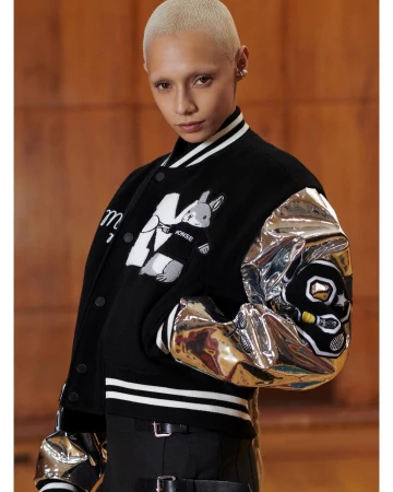 Model with bleached hair and eyebrows wearing a black varsity jacket featuring metallic silver sleeves and graphic appliqués.