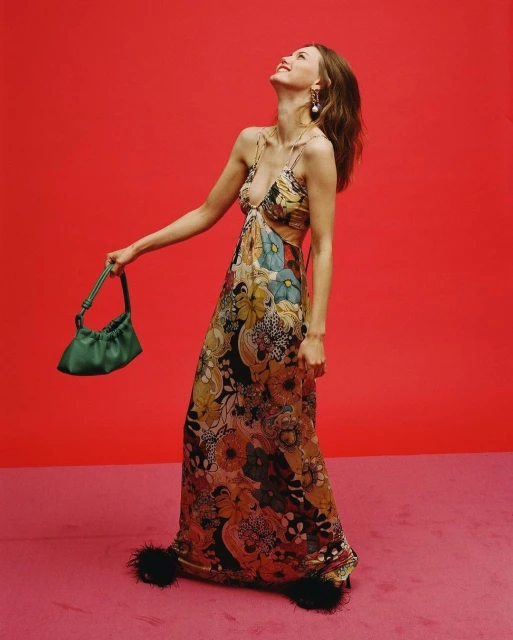 A young woman in a floral gown smiles, angled up against a red backdrop, holding a green purse with feather-trimmed heels in view.