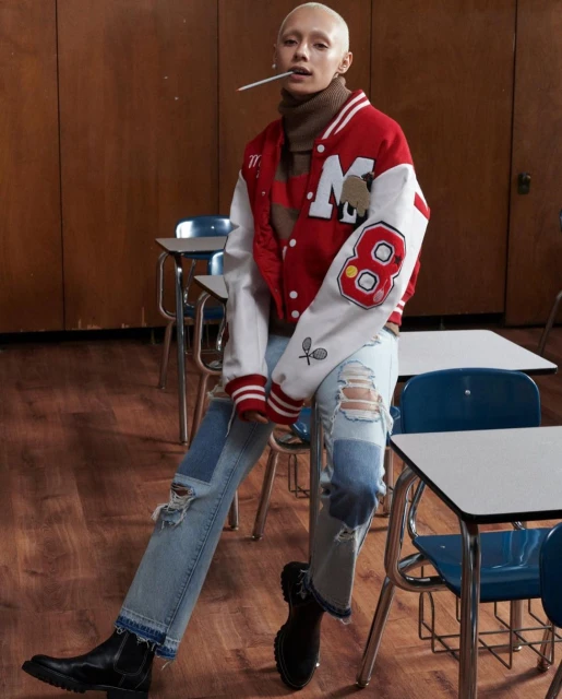 Fashion portrait of a short-haired adult wearing ripped jeans, varsity jacket, and boots, leaning on school desks in a classroom.