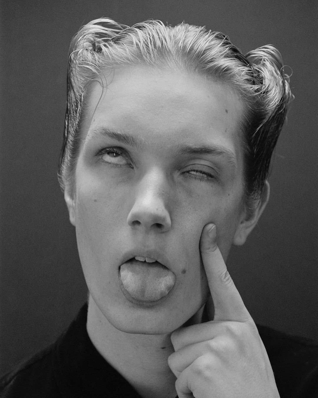 Close-up portrait of a young adult woman with stylized blonde hair, sticking out her tongue and winking in a black and white photograph.