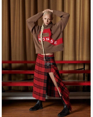 Model with shaved head and long braided hair wearing brown knit sweater and red plaid skirt standing indoors