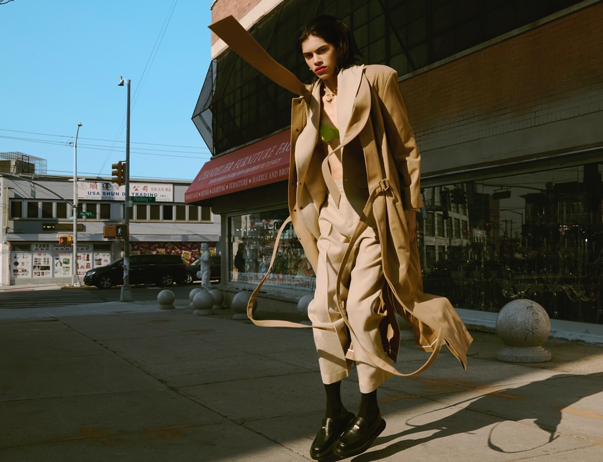 Fashion shot of a woman in a beige coat and pants standing on a city sidewalk in front of buildings with Chinese characters and furniture stores.