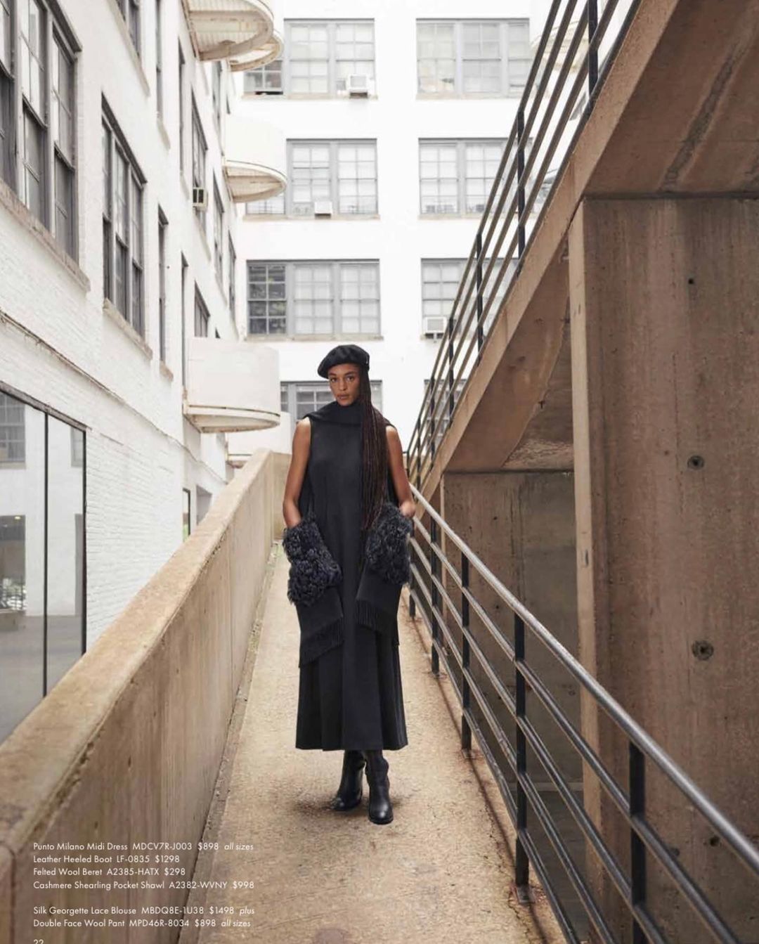 Woman in dark sleeveless dress wearing a beret standing on an outdoor ramp between white and concrete buildings.