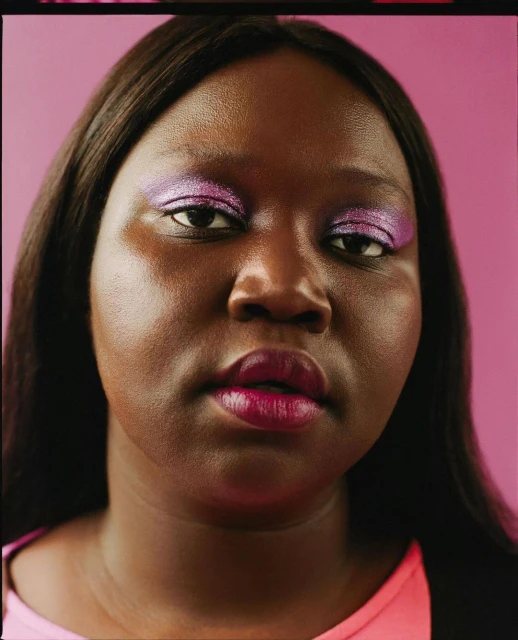 Portrait of a woman with purple eyeshadow and lipstick against a pink background, looking at the camera with neutral expression.