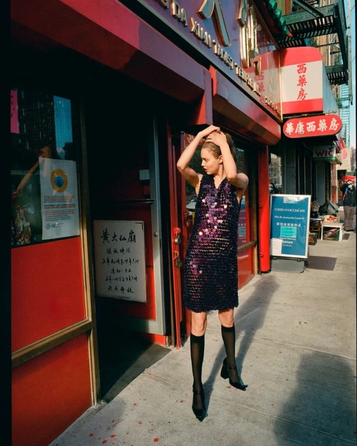 A fashion model poses in front of a red building wearing a sparkling black dress and black knee-high socks on a sunny city street.