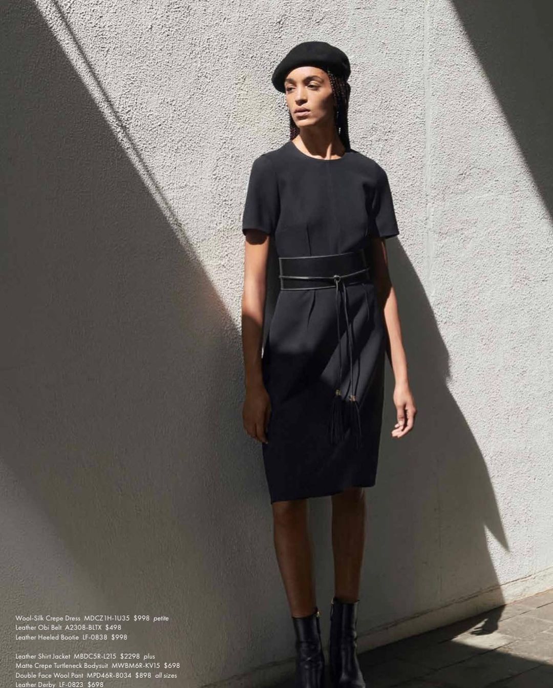 A woman poses in a black dress with a belt, black hat, and ankle boots against a textured white wall with light and shadow.