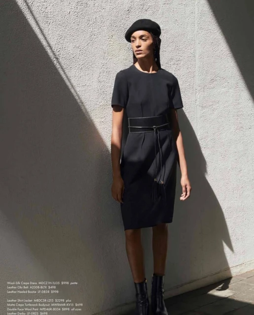 A woman poses in a black dress with a belt, black hat, and ankle boots against a textured white wall with light and shadow.
