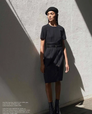 A woman poses in a black dress with a belt, black hat, and ankle boots against a textured white wall with light and shadow.