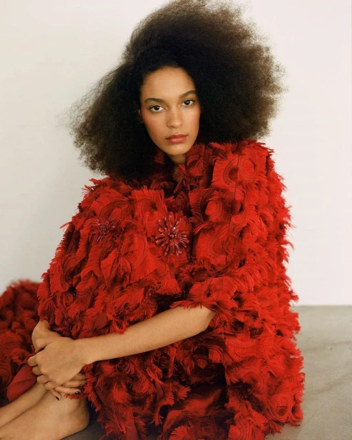 A woman with dark curly hair in a large red, textured coat poses indoors against a neutral background in a fashion editorial style.